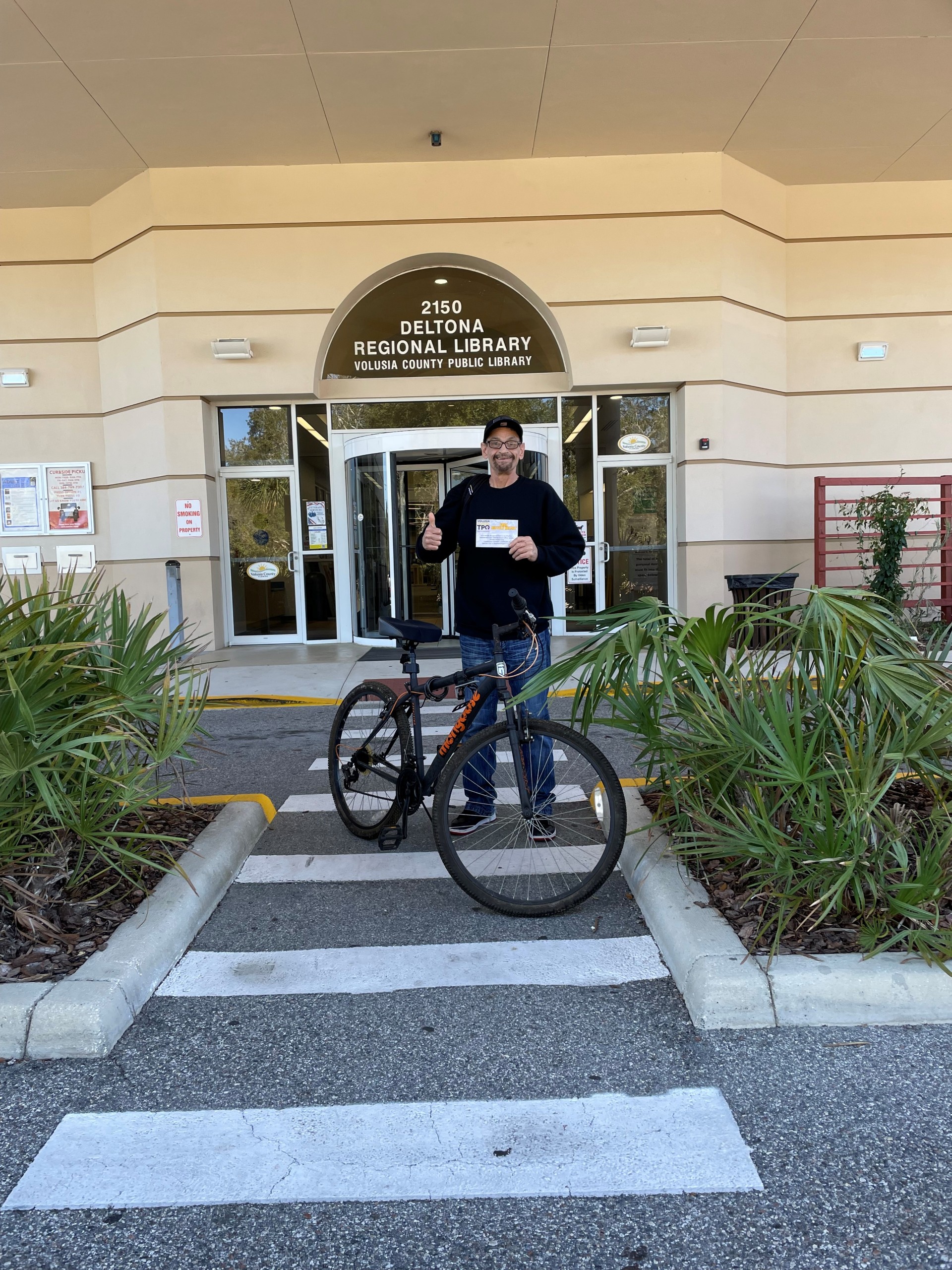 A person outside the Deltona Regional Library next to a bicycle holding a certificate and giving a thumbs up.